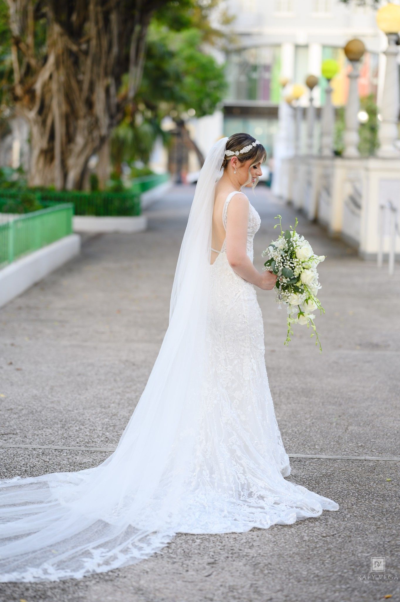 Bride in elegant lace mermaid wedding gown with long veil holds white bouquet outdoors on a sunny day.