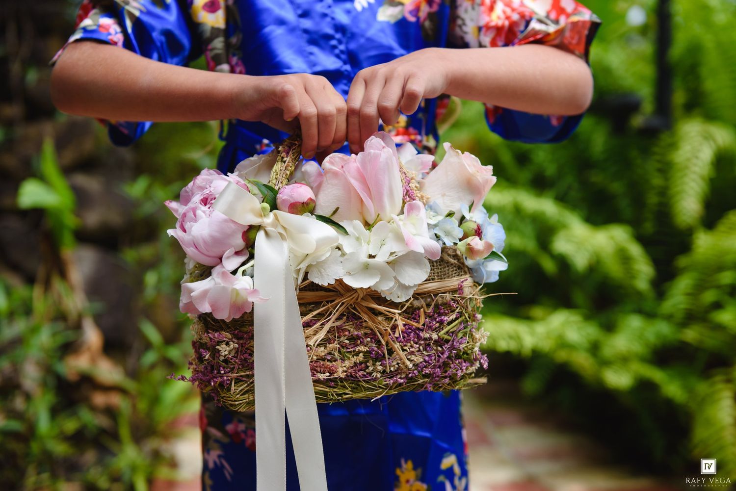 A flower girl in a blue floral dress holds a rustic basket filled with pink and white flowers tied with a white ribbon.