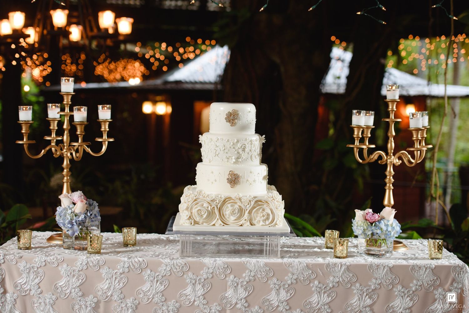 Elegant white five-tier wedding cake with floral details on lace-covered table flanked by gold candelabras in garden setting.