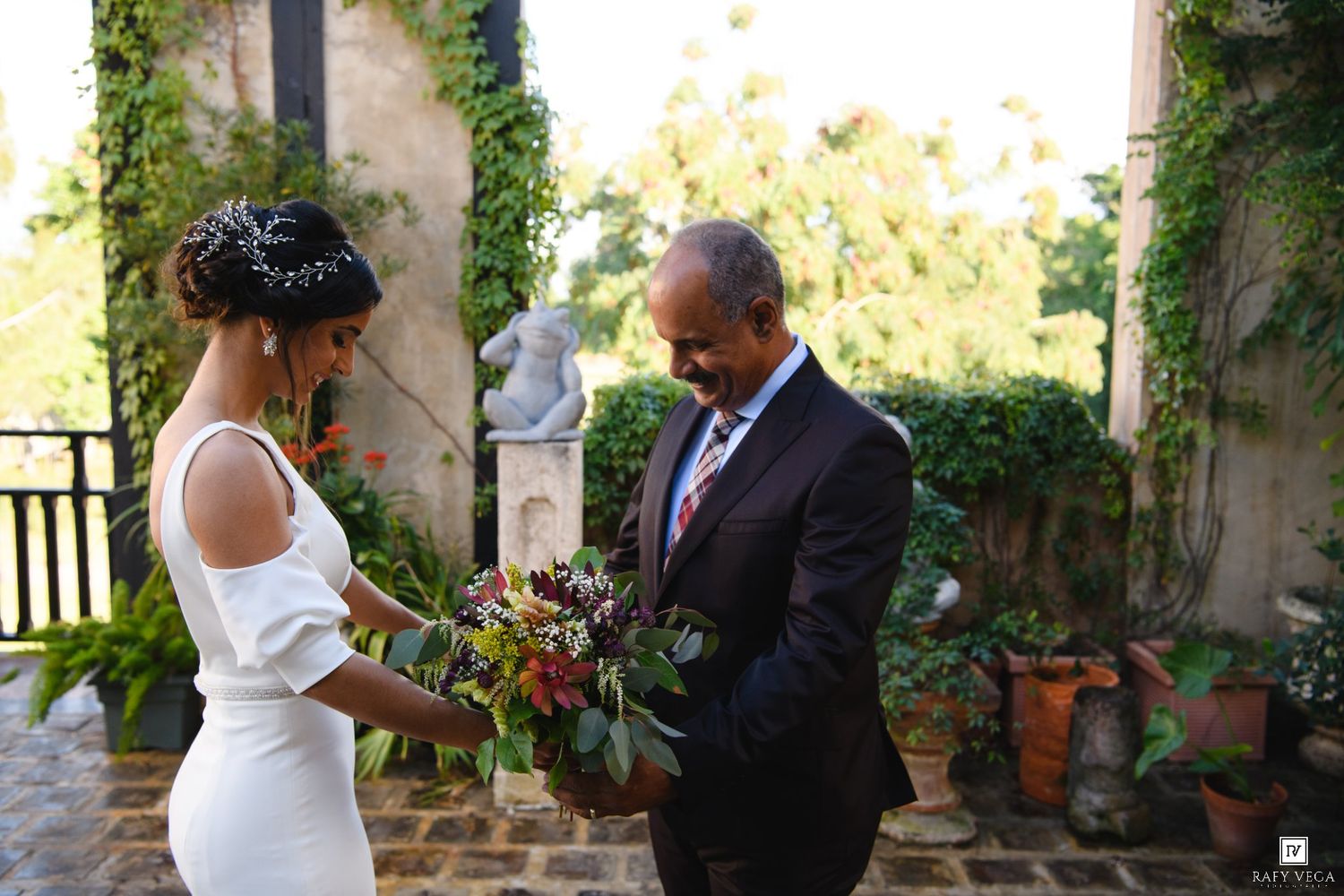 Bride in white gown holding tropical bouquet with groom in suit at elegant ivy-covered outdoor venue.