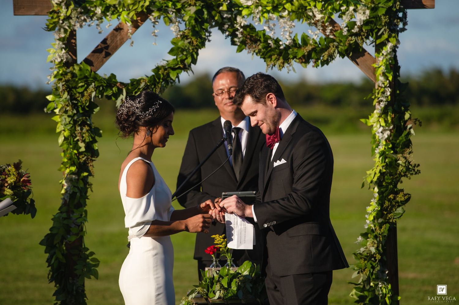 Groom places wedding ring on bride's finger during outdoor ceremony under floral arch with officiant.