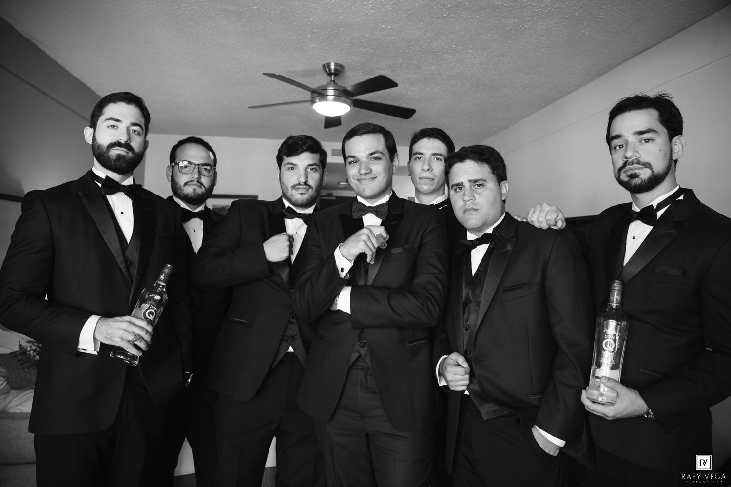Black and white photo of groomsmen in tuxedos holding champagne bottles, posing together before a wedding ceremony.