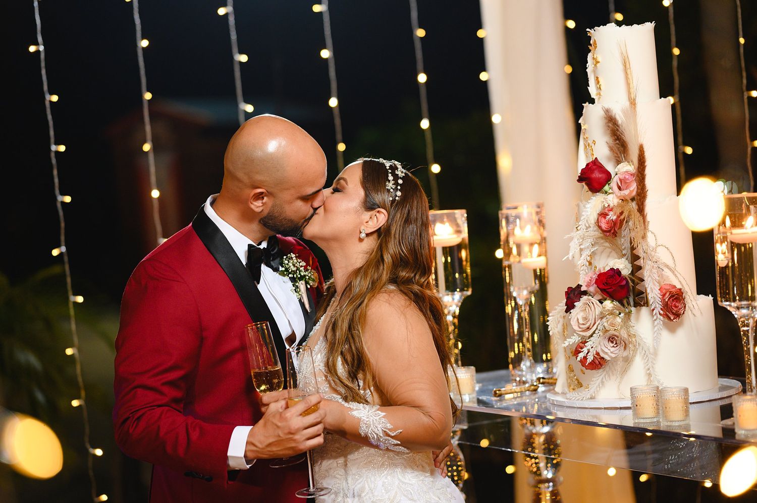 Bride and groom share a kiss beside a floral wedding cake at an elegant evening reception with fairy lights.