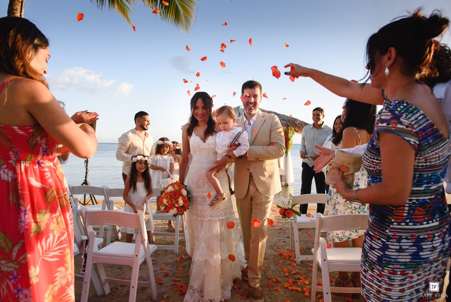 Couple walks down beach wedding aisle as guests toss red and orange flower petals under a tropical sky.