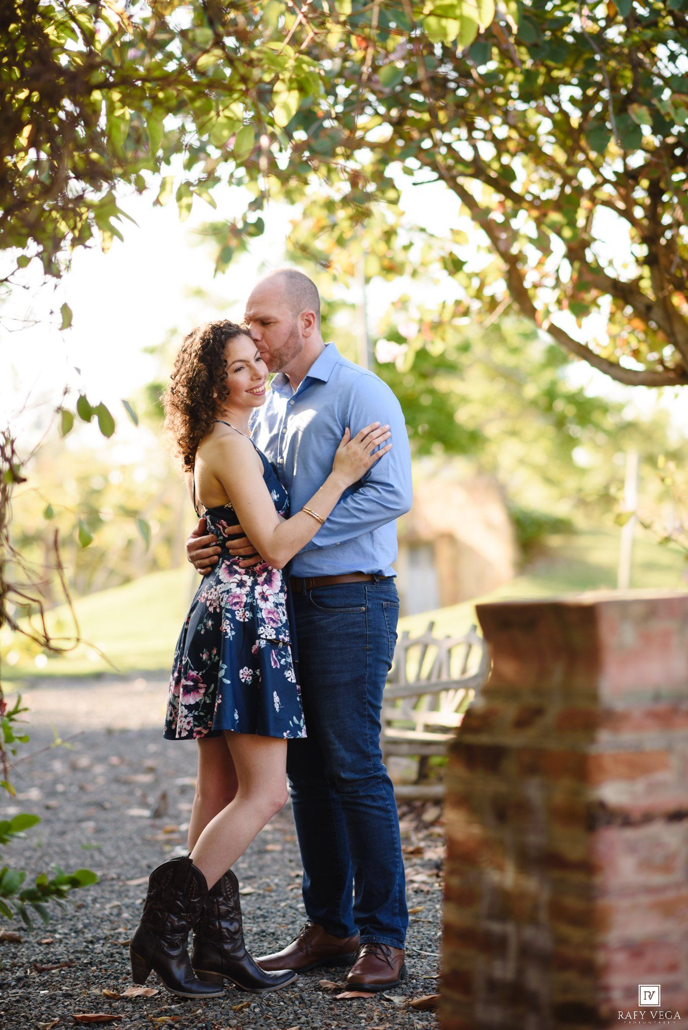 Couple embracing under sunlit trees in a garden, woman in floral dress and boots, man in blue shirt and jeans.