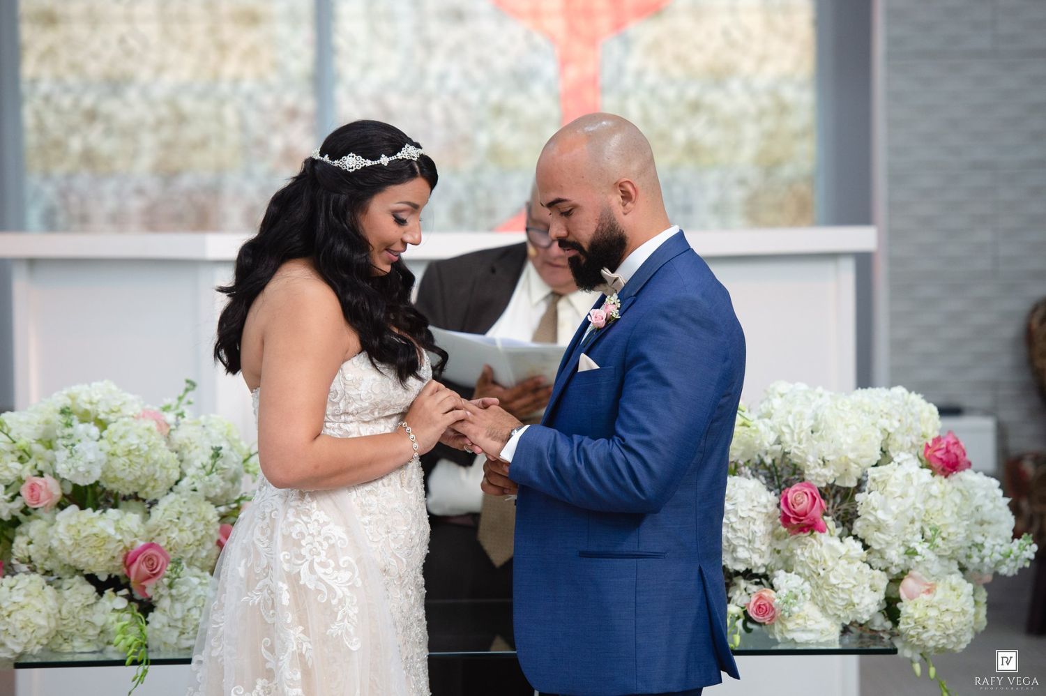 Bride and groom exchanging rings at wedding ceremony with white floral arrangements and stained glass backdrop.