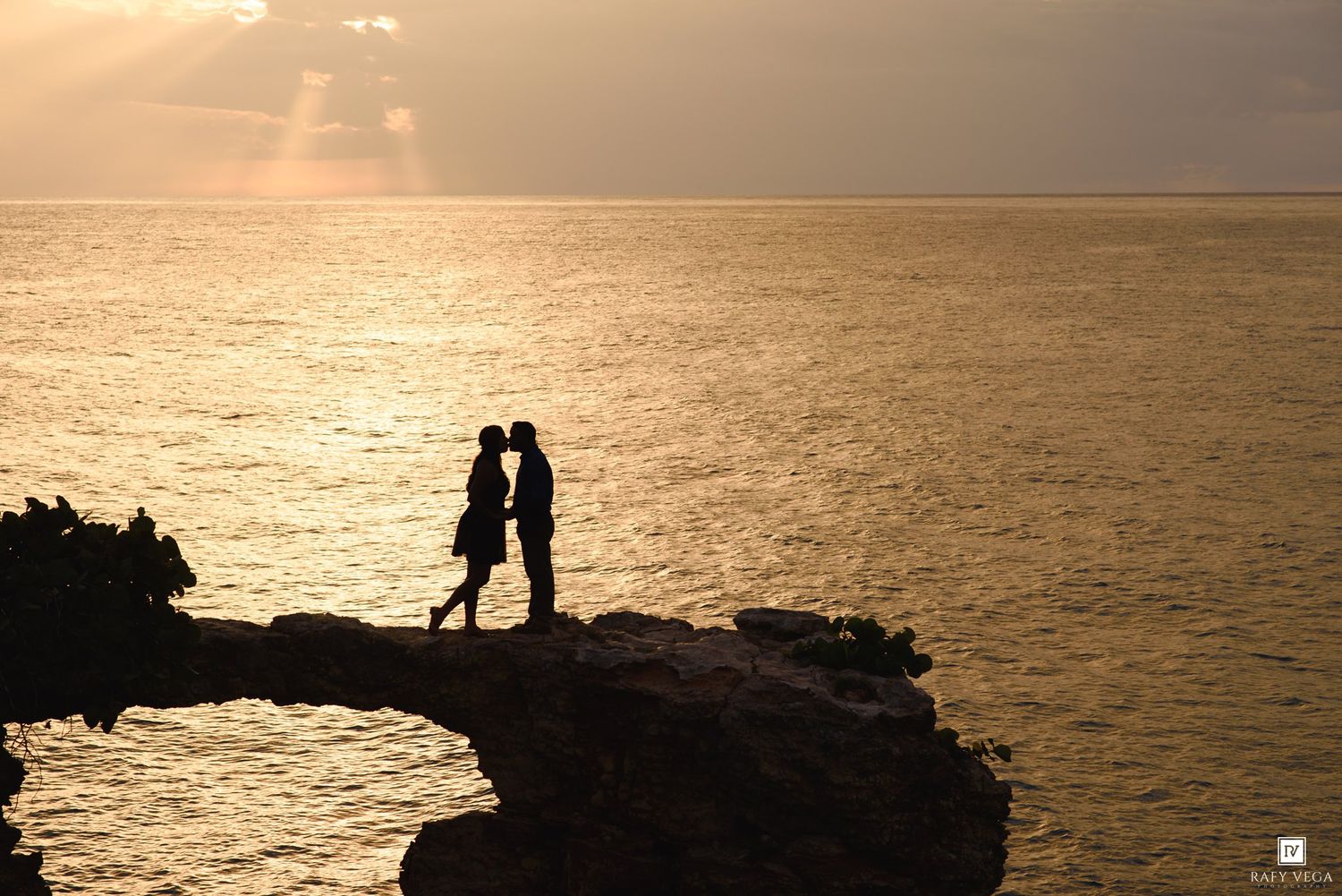 Silhouette of a couple kissing on rocky cliffs at golden sunset over the ocean.