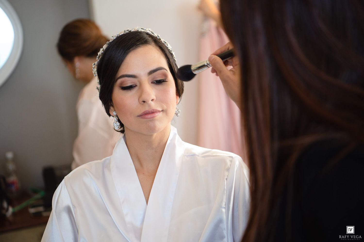 A bride in a white robe gets makeup applied with a brush during wedding preparations.