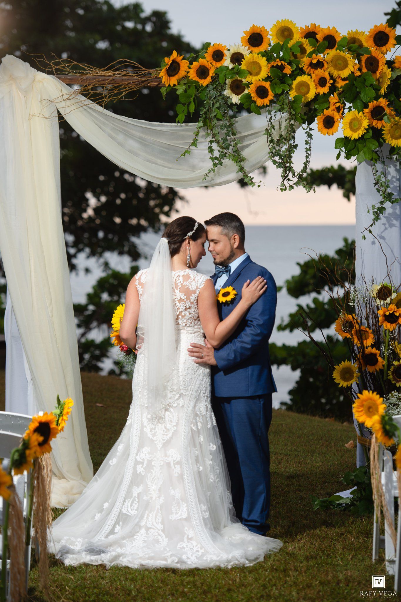 Bride and groom embrace under a sunflower-adorned arch with white draping at an outdoor wedding ceremony.