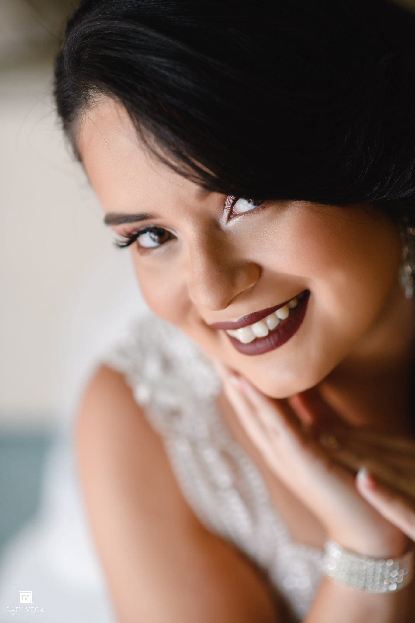 Smiling bride in beaded white gown with dark updo and berry lips, posing elegantly for a close-up wedding portrait.