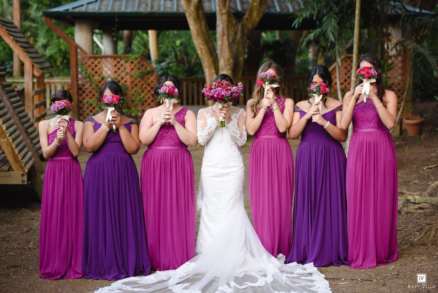 Bride in white gown poses with bridesmaids in purple and fuchsia dresses holding bouquets covering their faces outdoors.
