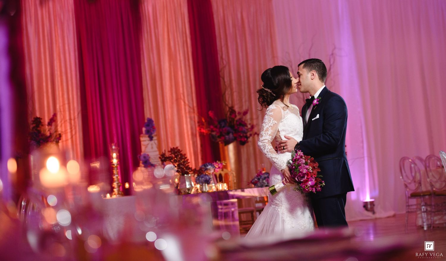 Bride and groom share a romantic kiss at a candlelit wedding reception with pink and red floral decor.
