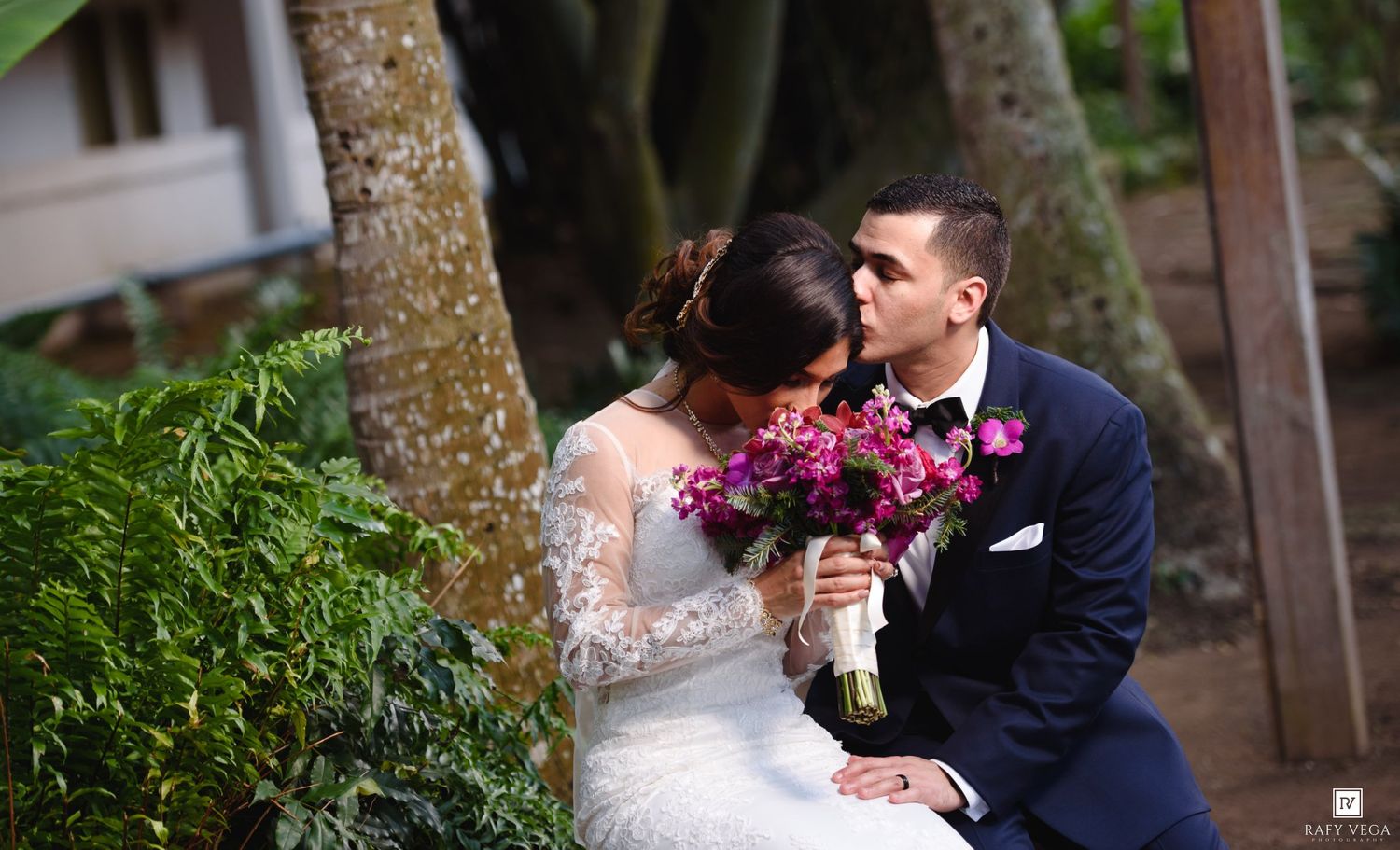 Groom kisses bride holding purple bouquet among tropical trees in lush garden wedding portrait.