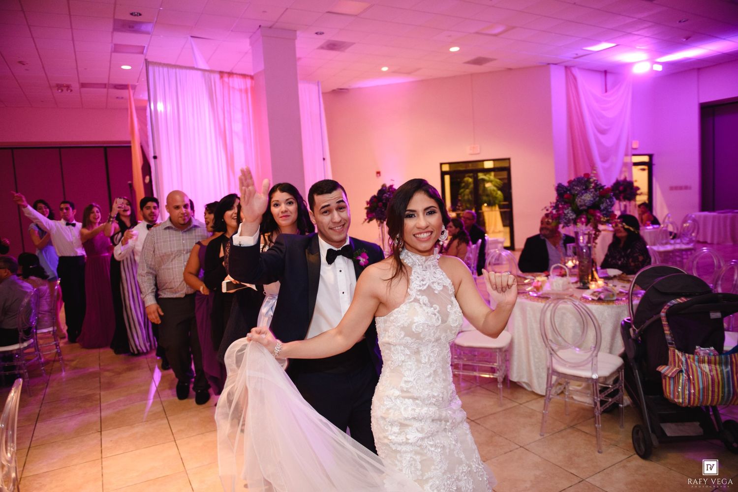 Bride and groom share first dance at elegant wedding reception with pink uplighting and guests celebrating in background.