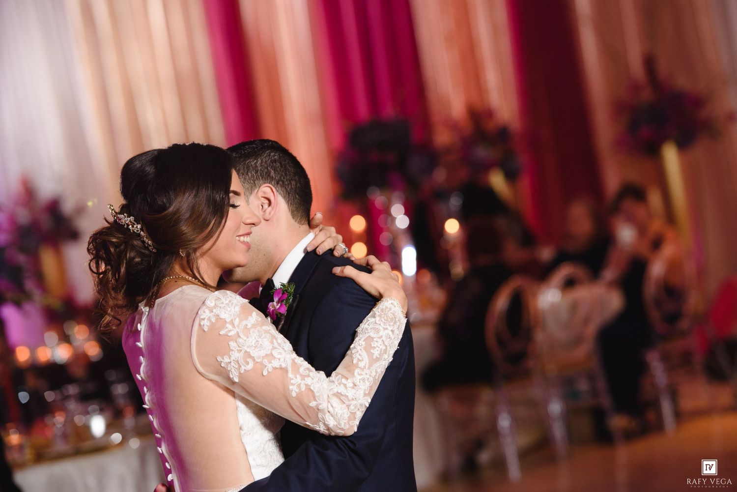Bride and groom share first dance kiss at elegant wedding reception with pink and gold floral backdrop.