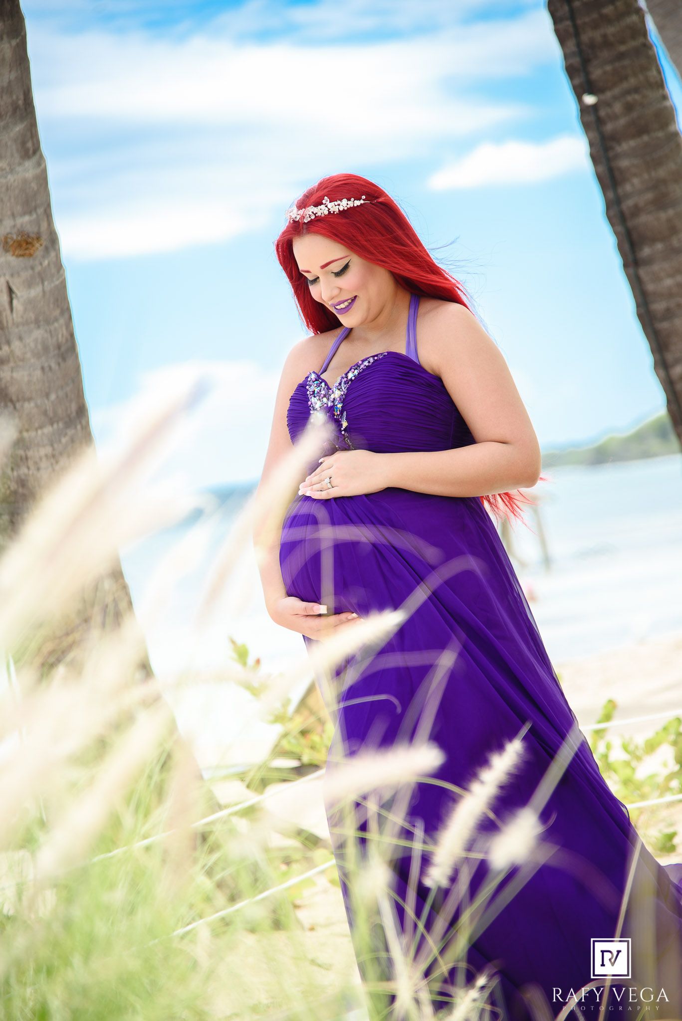 Smiling pregnant woman in purple dress with red hair cradling bump by ocean.