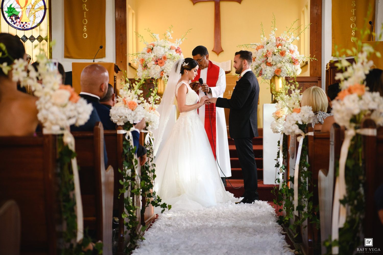 Bride and groom exchange vows at a church altar adorned with white and peach floral arrangements during a wedding ceremony.