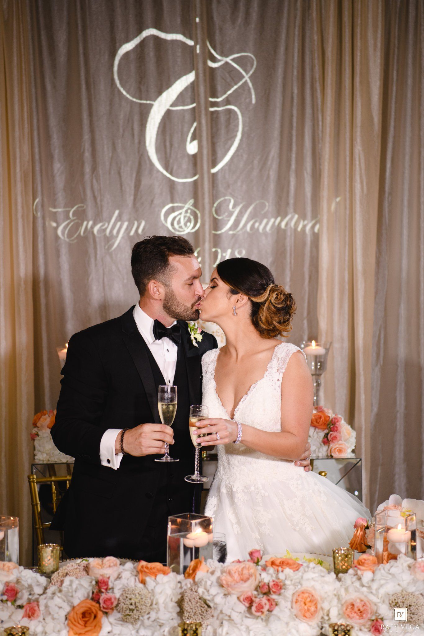 Bride and groom share a kiss at their wedding reception sweetheart table adorned with peach and white floral arrangements.