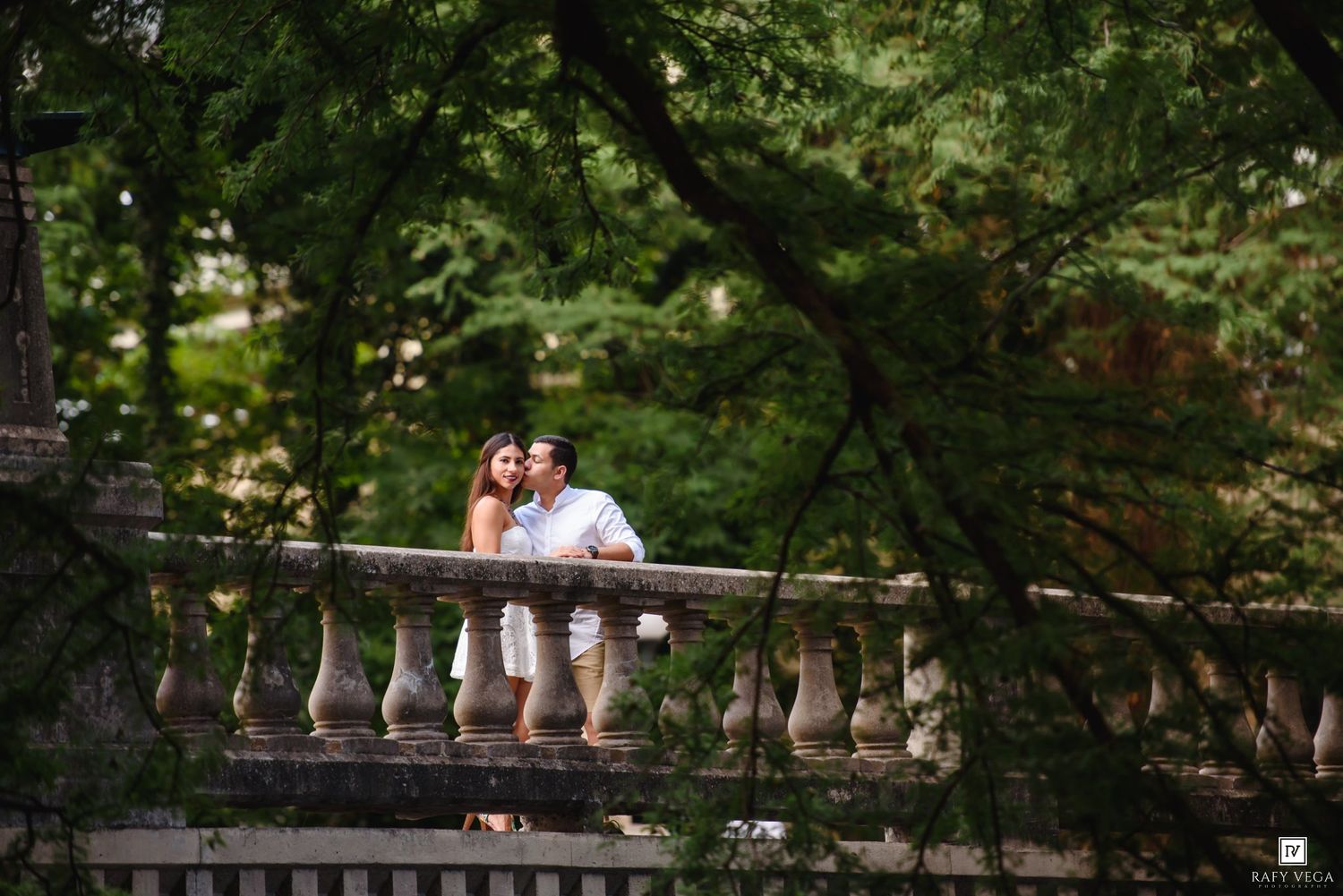 Couple sharing a romantic moment on a stone balustrade bridge surrounded by lush green trees.