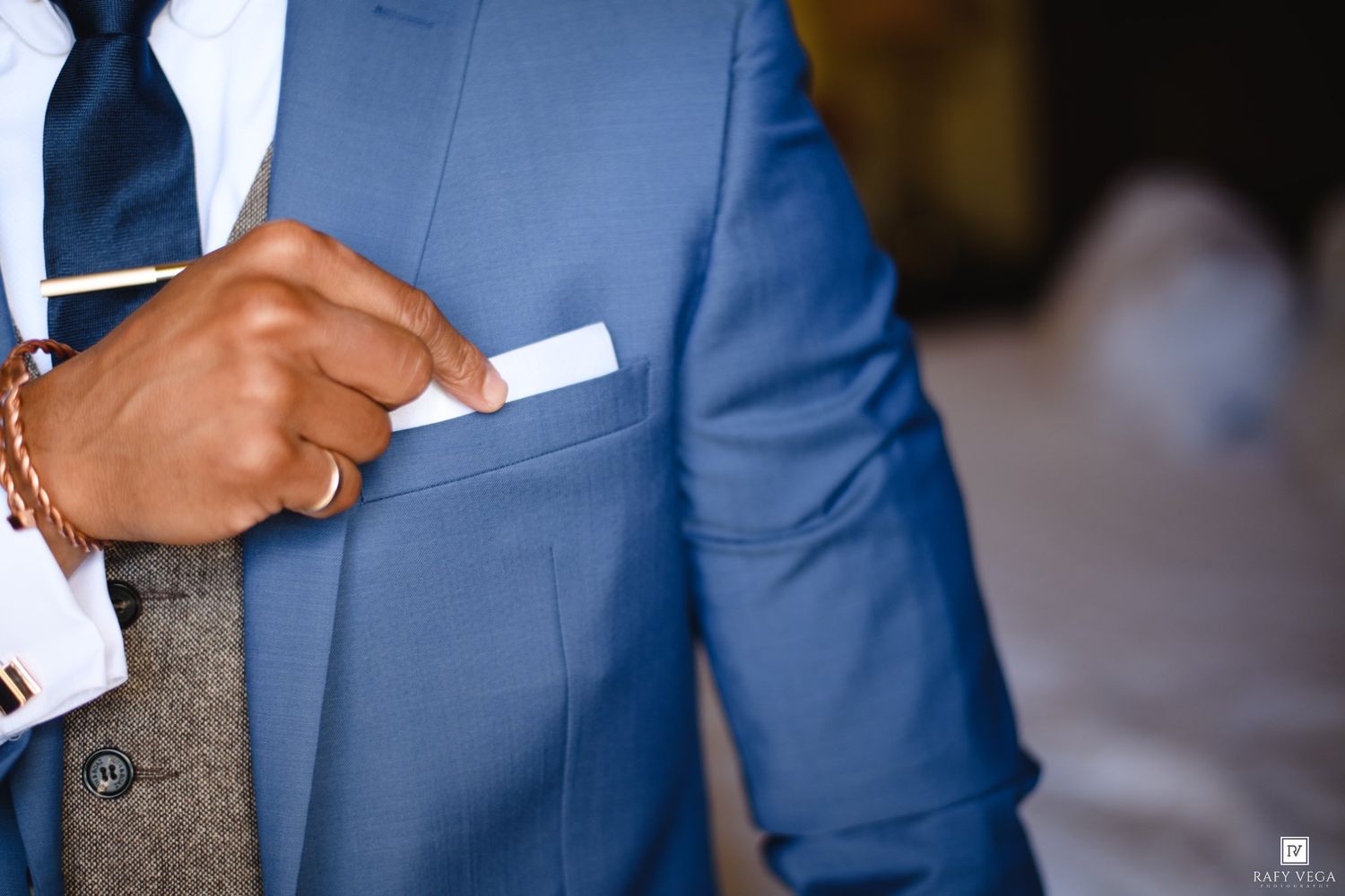 Man in blue suit adjusting white pocket square, wearing cufflinks and navy tie.
