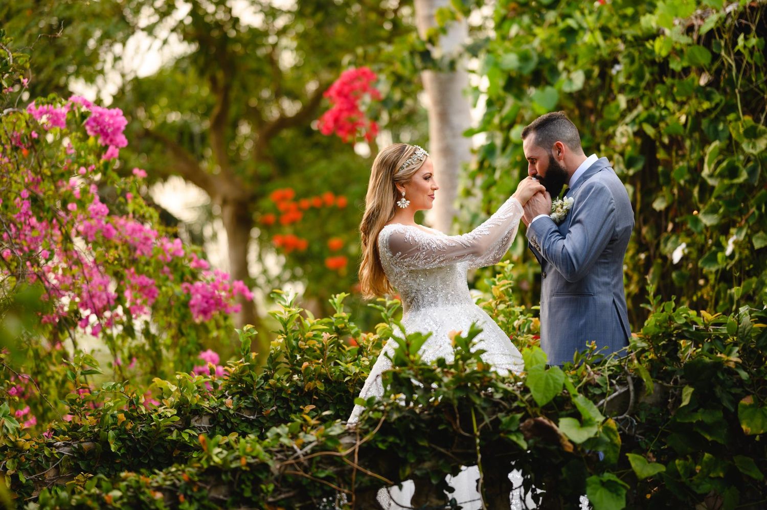 Groom kissing bride's hand among lush greenery and vibrant pink flowers in a romantic outdoor wedding setting.