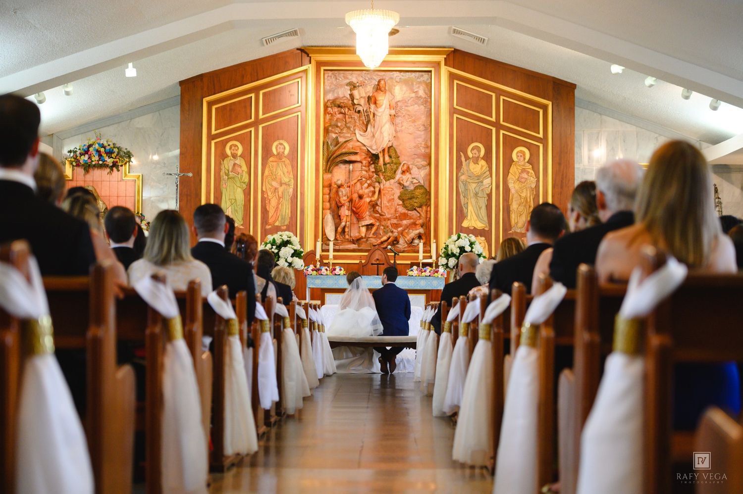 Elegant church wedding ceremony with guests seated in pews, couple at altar beneath ornate religious artwork.