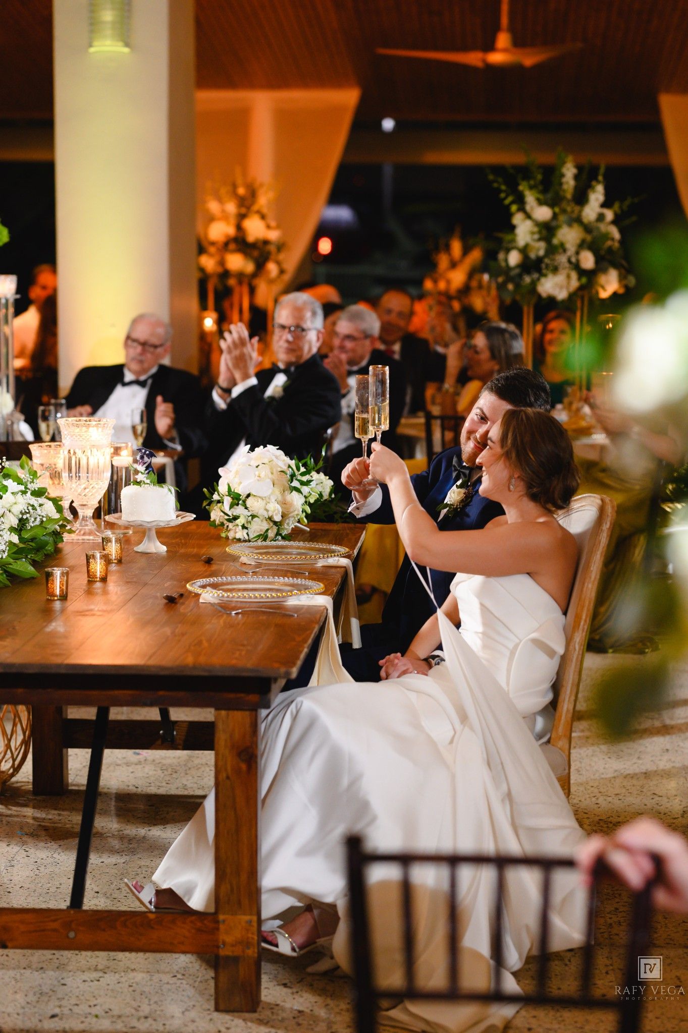Bride and groom toasting champagne at wedding reception table with floral arrangements and guests celebrating in background.