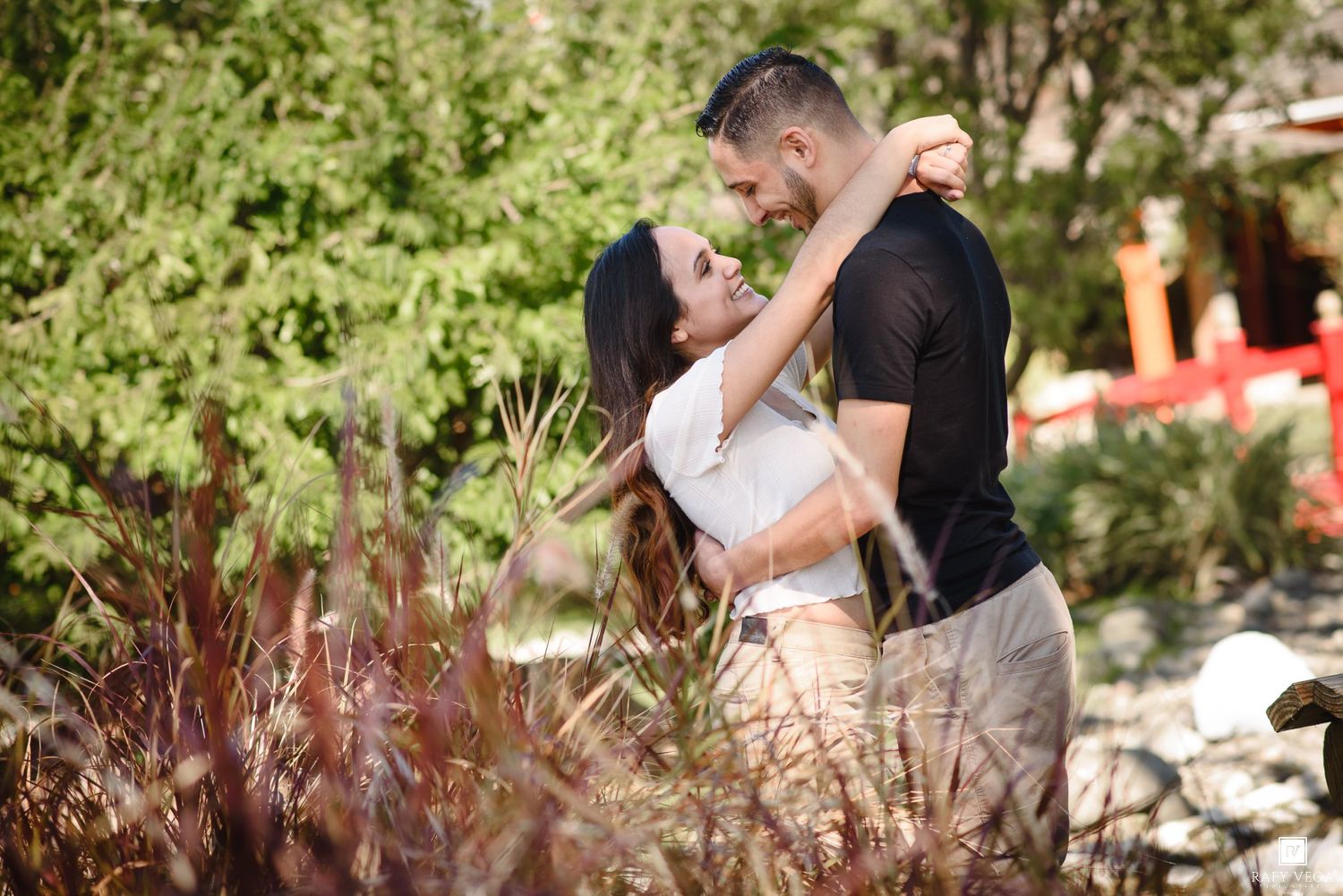 Couple embracing romantically outdoors among lush greenery and red foliage on a sunny day.