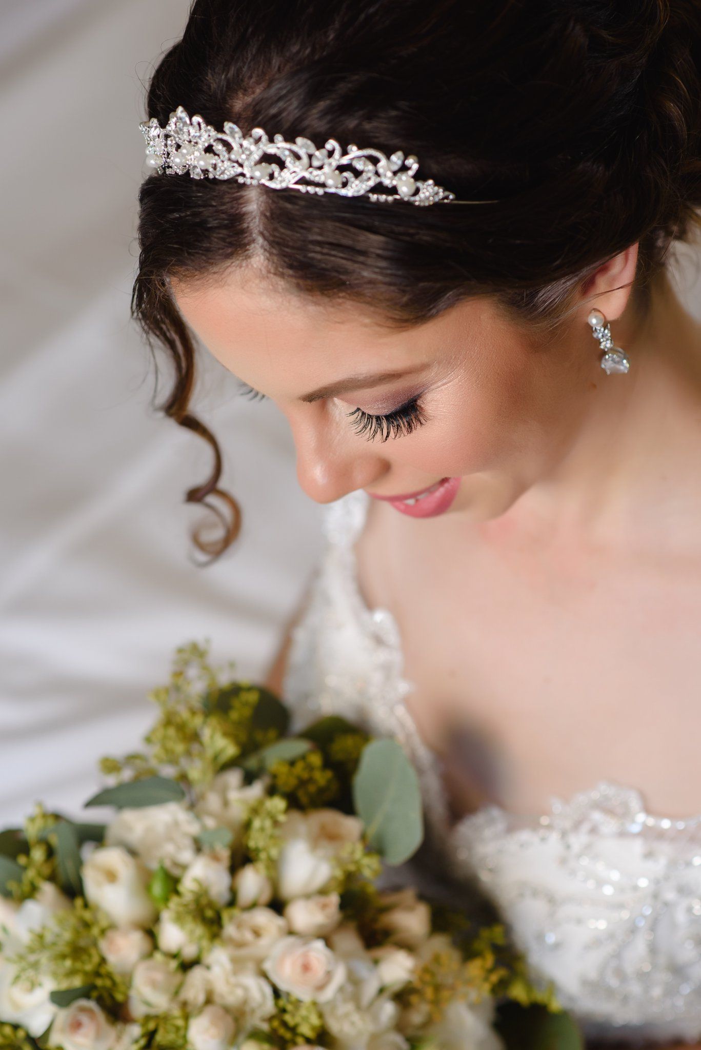 Elegant bride wearing a crystal tiara and drop earrings, looking down at her white and green wedding bouquet.