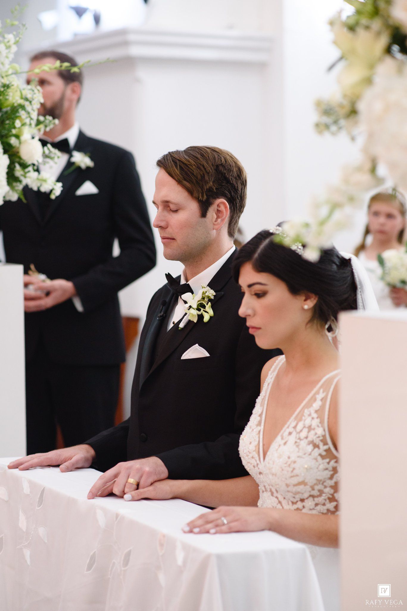 Bride and groom sit together at altar during elegant church wedding ceremony, groom in black tuxedo with floral boutonniere.