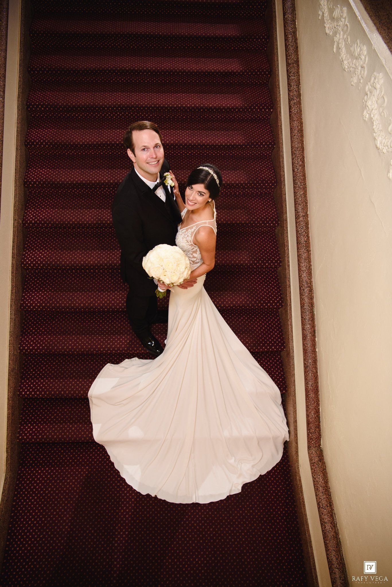 Aerial view of bride in white gown holding bouquet and groom in tuxedo posing on red carpeted staircase.