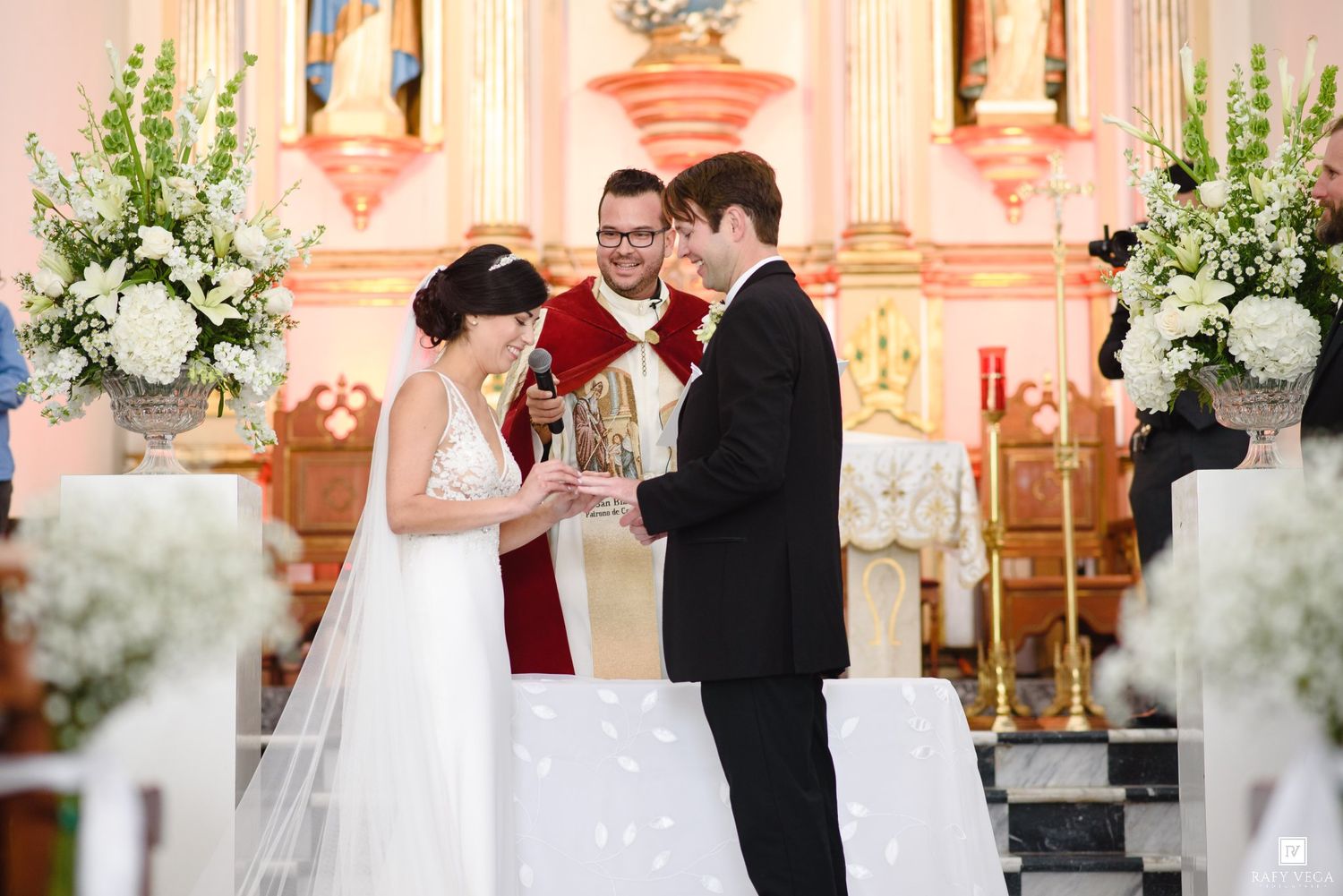 Bride and groom exchange vows at a church altar adorned with white floral arrangements during their wedding ceremony.