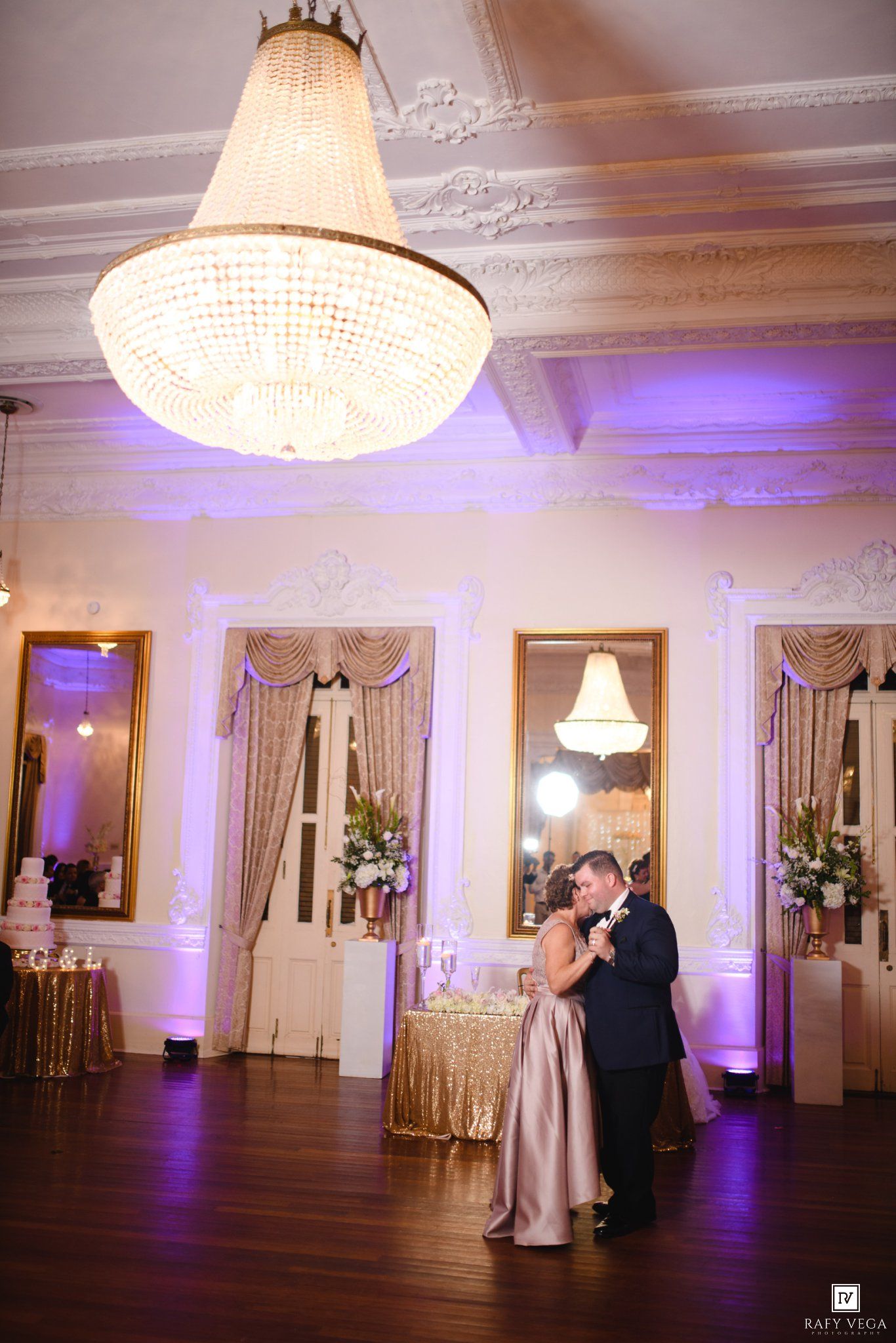 Bride and groom share first dance under crystal chandelier in elegant ballroom with purple uplighting.