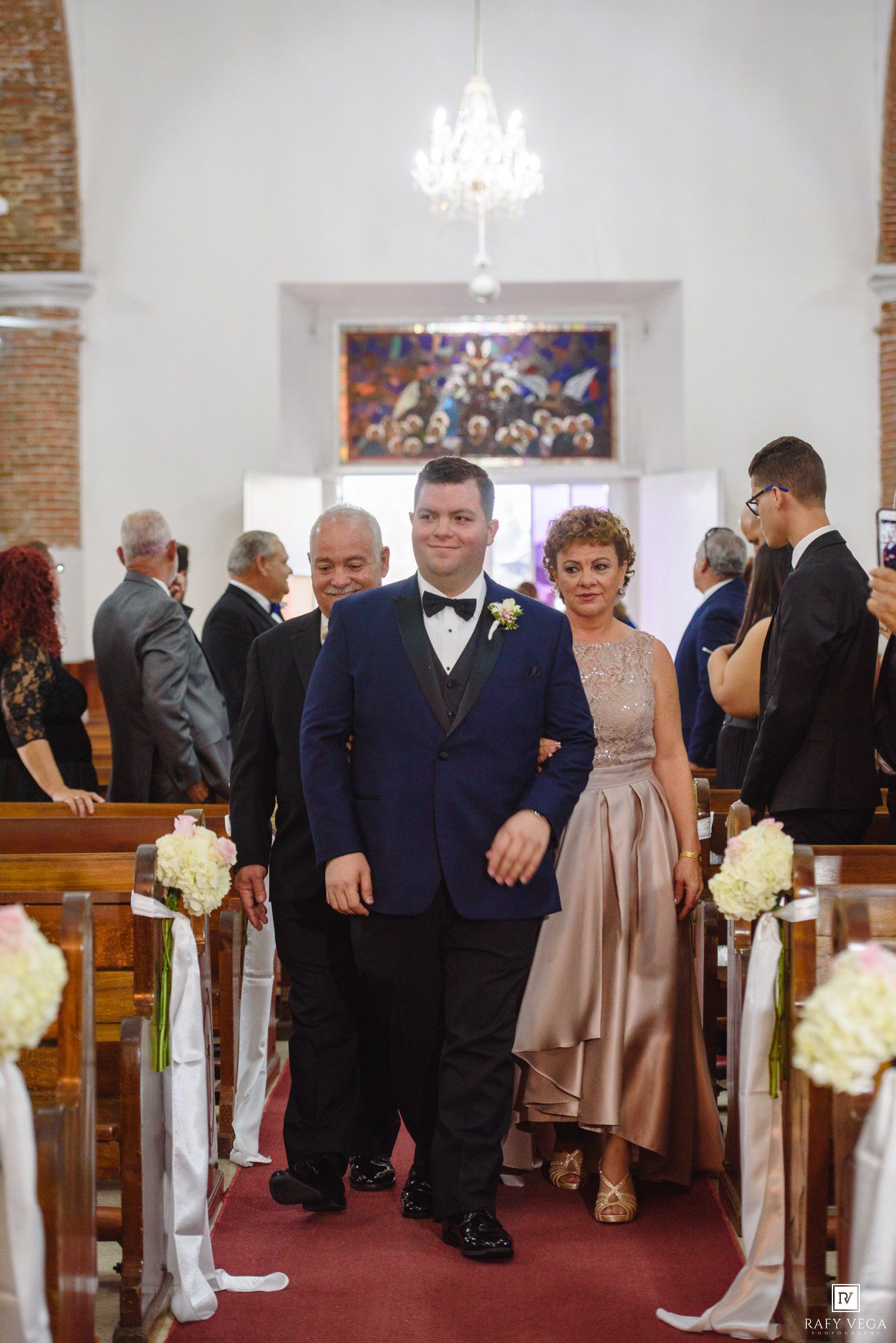 Groom in navy tuxedo walks down church aisle with mother in gold dress during wedding ceremony.