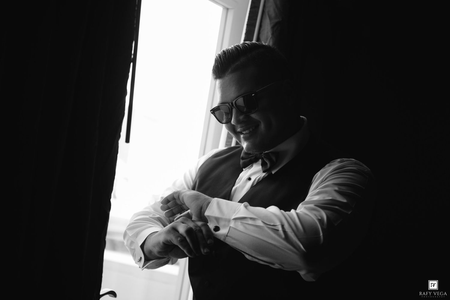 Black and white photo of a well-dressed man in vest and bow tie adjusting his cufflinks by a window.