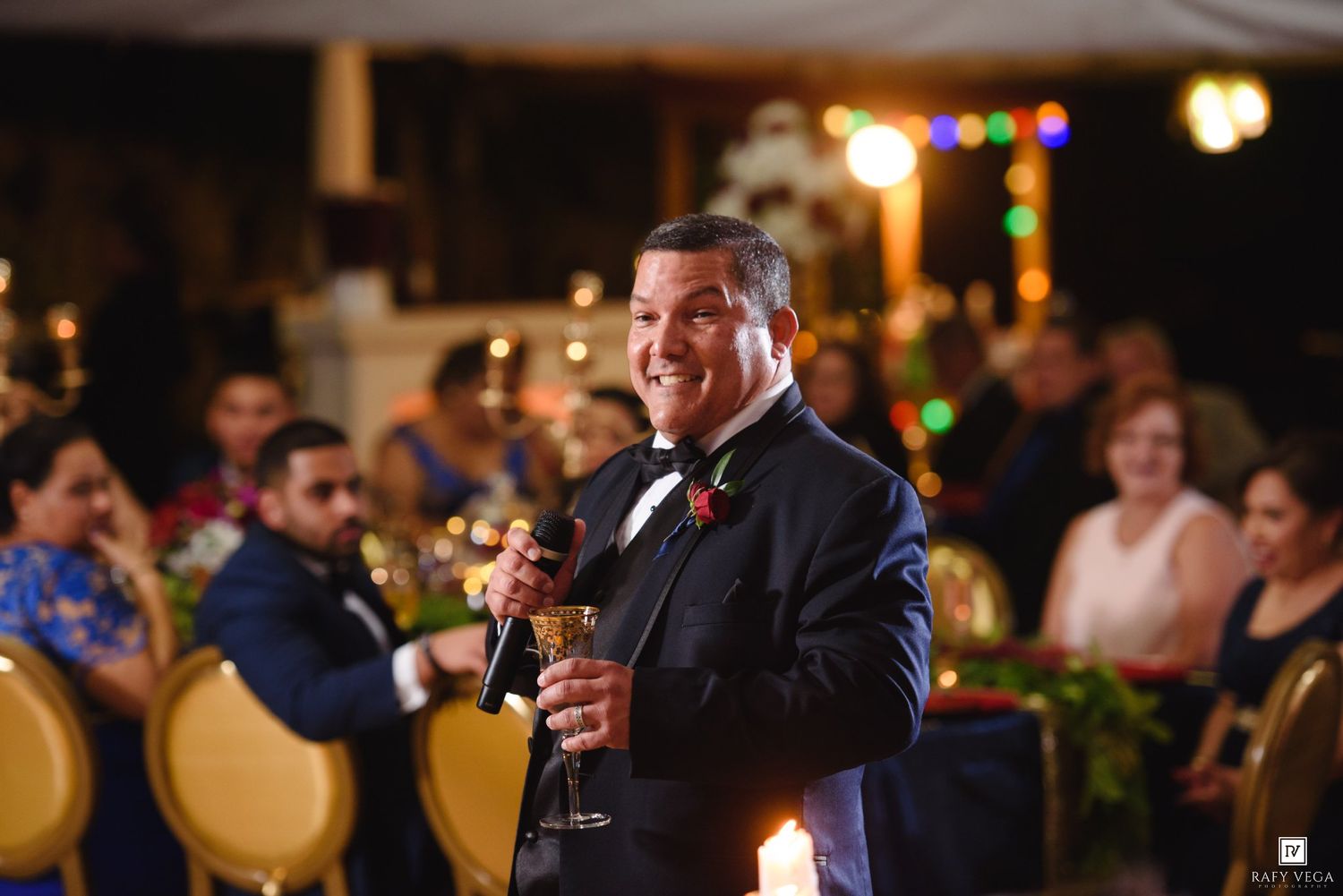 Man in tuxedo giving a toast at wedding reception holding champagne glass with guests seated in background.