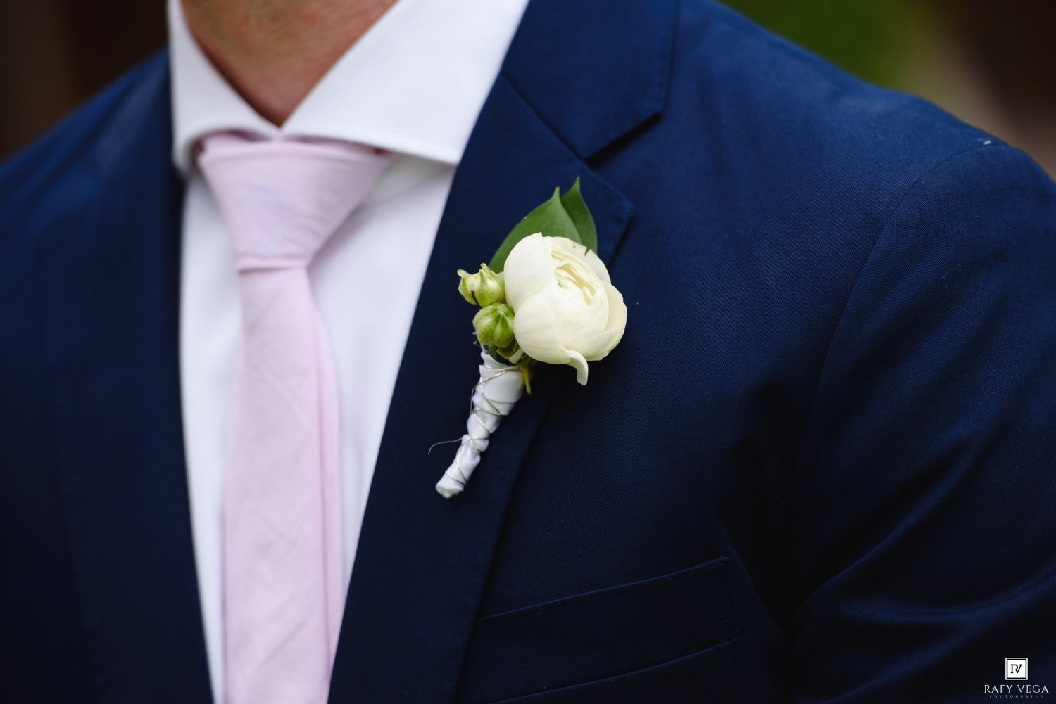 Groom wearing navy blue suit with white rose boutonniere and pink tie at wedding.