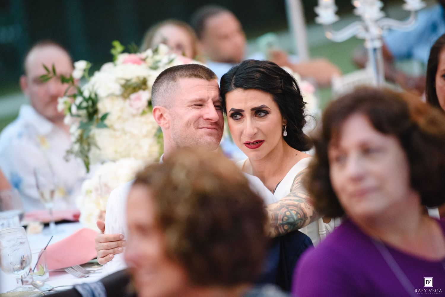 Bride and groom sitting together at wedding reception dinner table, surrounded by guests and floral centerpieces.