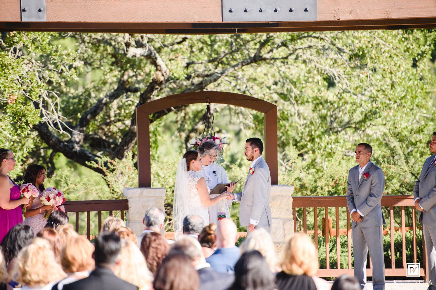 Couple shares vows at outdoor wedding ceremony under rustic pergola with guests and bridal party.