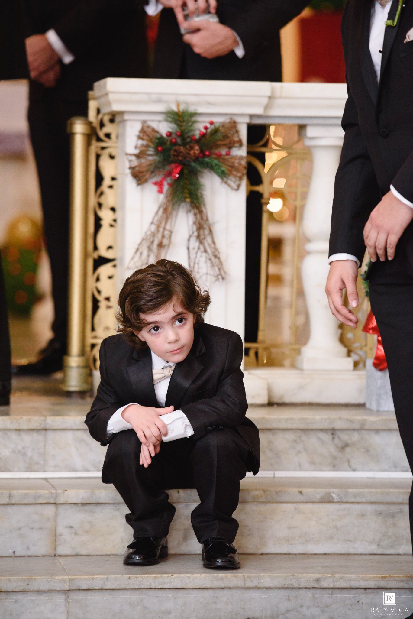 Young ring bearer in black tuxedo crouching on marble church steps during a Christmas wedding ceremony.