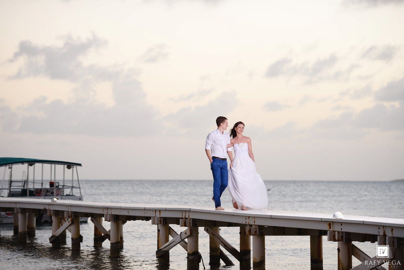 Couple in wedding attire standing on a wooden pier over calm ocean waters at sunset.