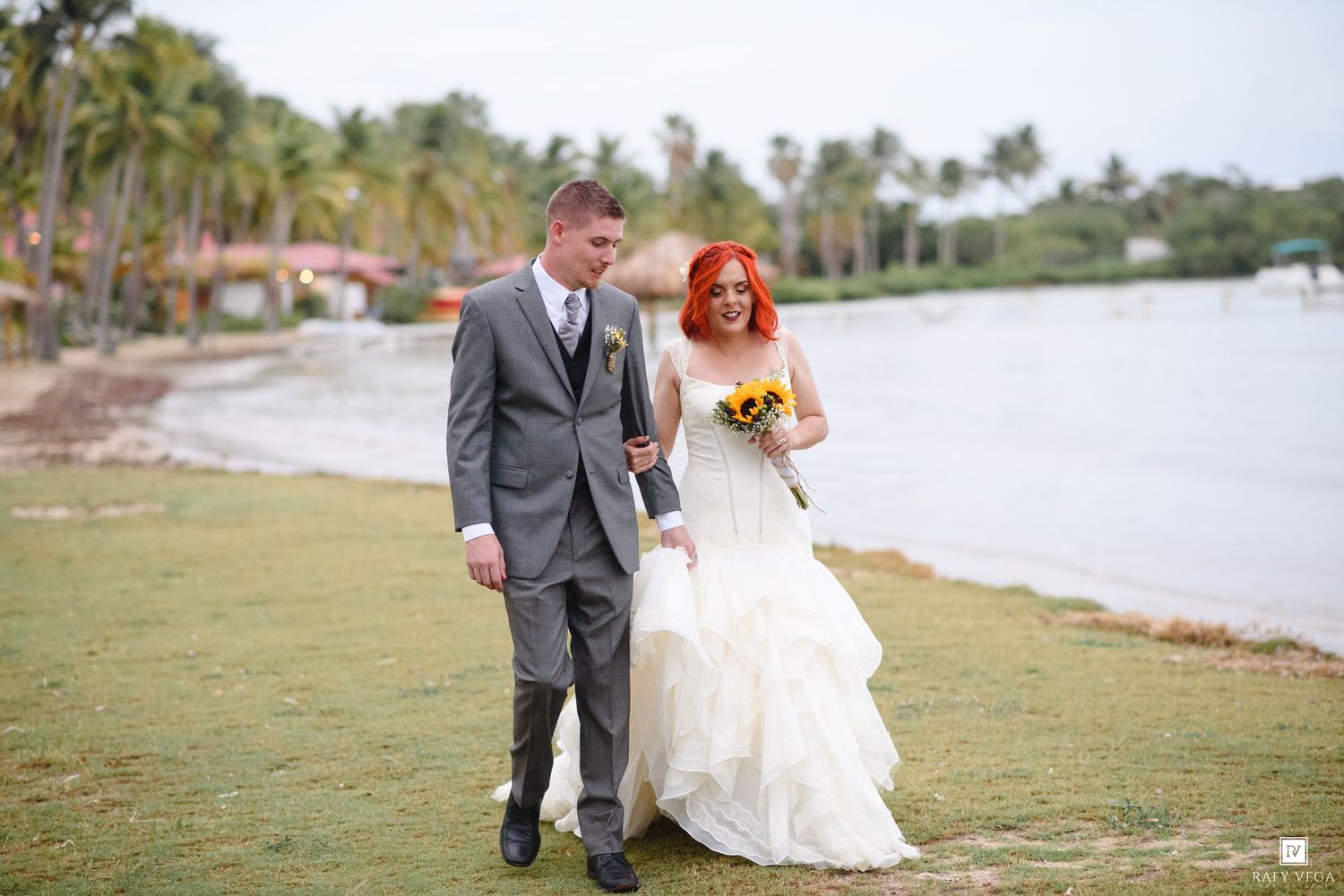 Bride in white gown with red hair holding bouquet walks with groom in gray suit near tropical beach.