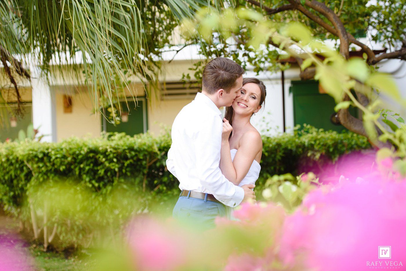 A couple shares a romantic embrace outdoors surrounded by lush green hedges and vibrant pink flowers.