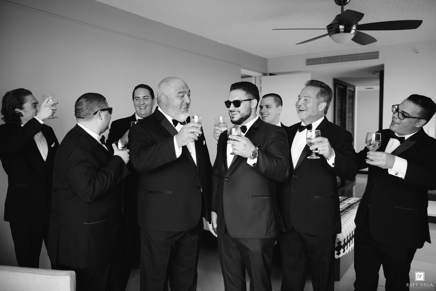 Groomsmen in tuxedos toasting with champagne glasses in a black and white wedding photo.