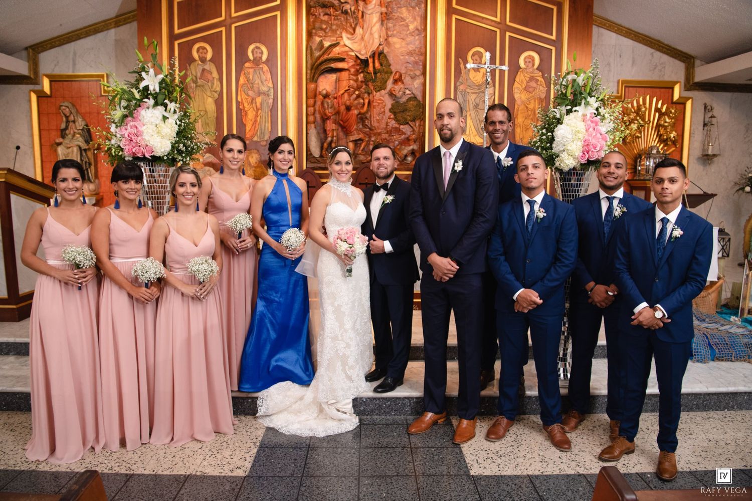 Wedding party poses in church with bridesmaids in pink gowns and groomsmen in navy suits flanking bride and groom.