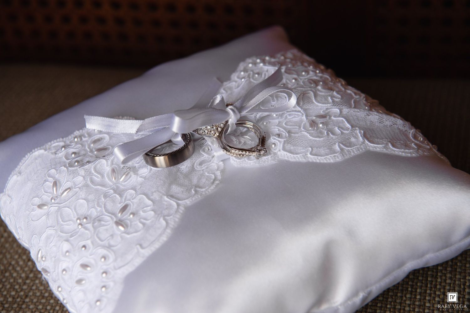 White satin wedding ring pillow with lace embroidery, ribbon bow, and two gold bands displayed elegantly.