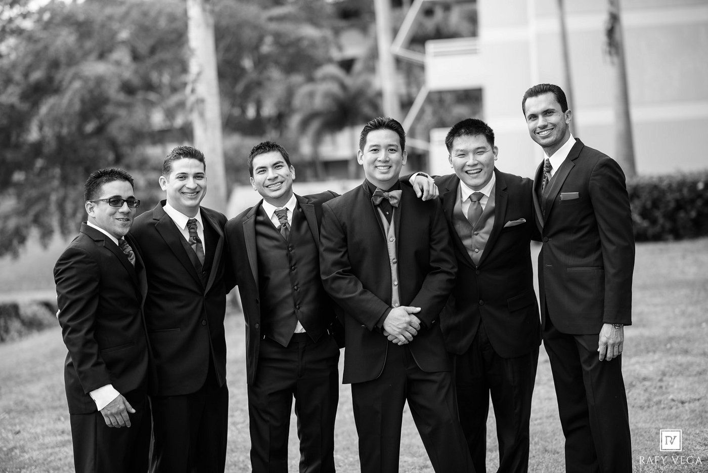 Black and white photo of six groomsmen in dark suits smiling together outdoors at a wedding.