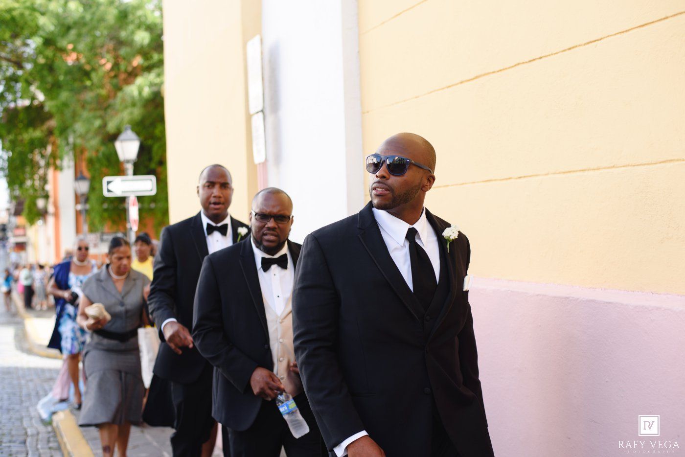 Groomsmen in black tuxedos walking confidently down a sunlit street during a wedding procession.