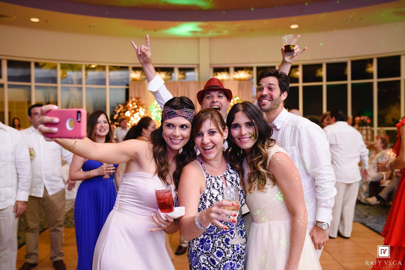 Group of friends taking a selfie at a festive party, wearing white and colorful outfits in a bright event venue.
