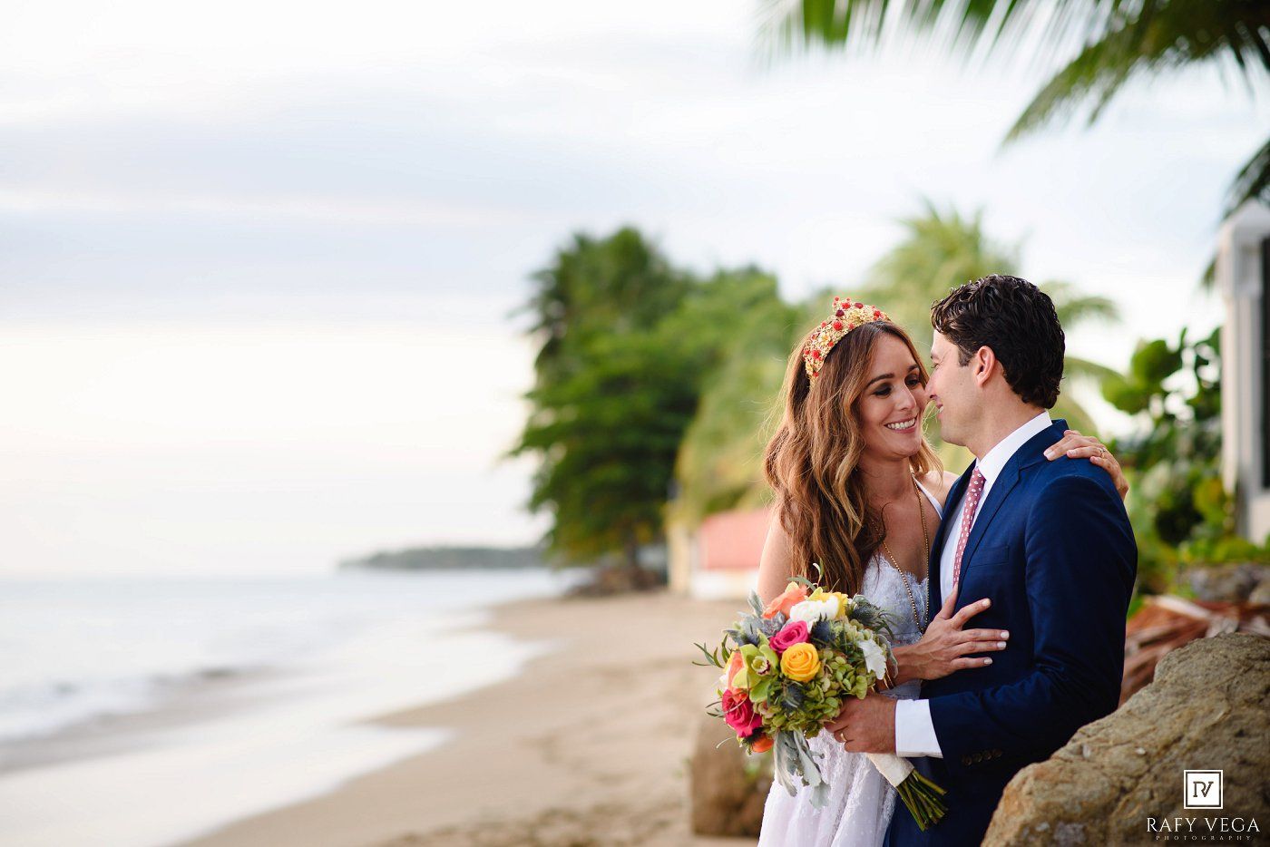 Bride and groom share a romantic moment on a tropical beach, with lush palms and ocean waves in the background.